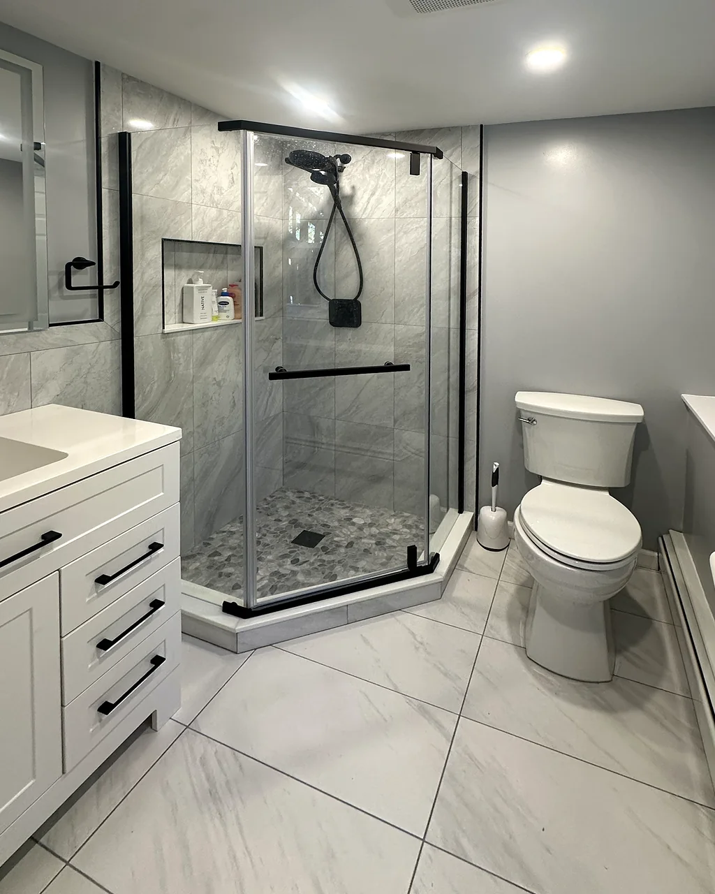 Remodeled bathroom with a neo-angle glass shower, marble-look tile, a pebble shower floor and a white shaker vanity