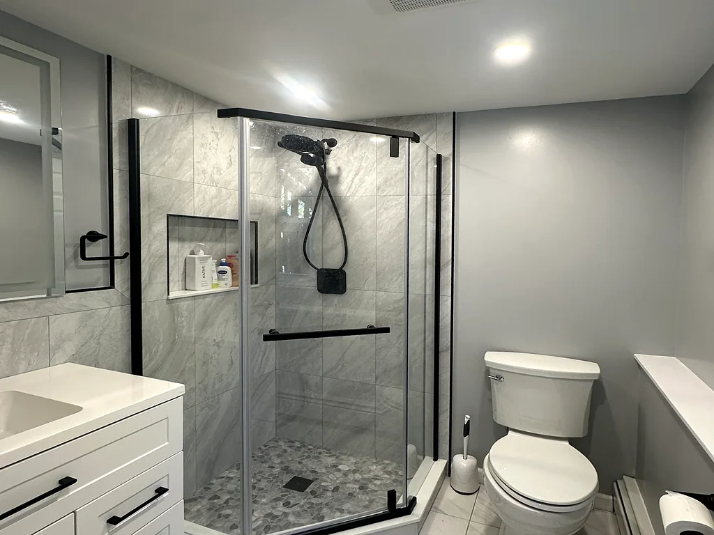 Remodeled bathroom with a neo-angle glass shower, marble-look tile, a pebble shower floor and a white shaker vanity