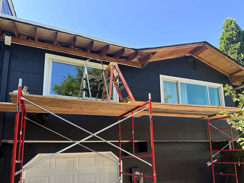 Scaffolding along a two-story house with the soffit opened up to the rafter tails and new fascia going on at the gable