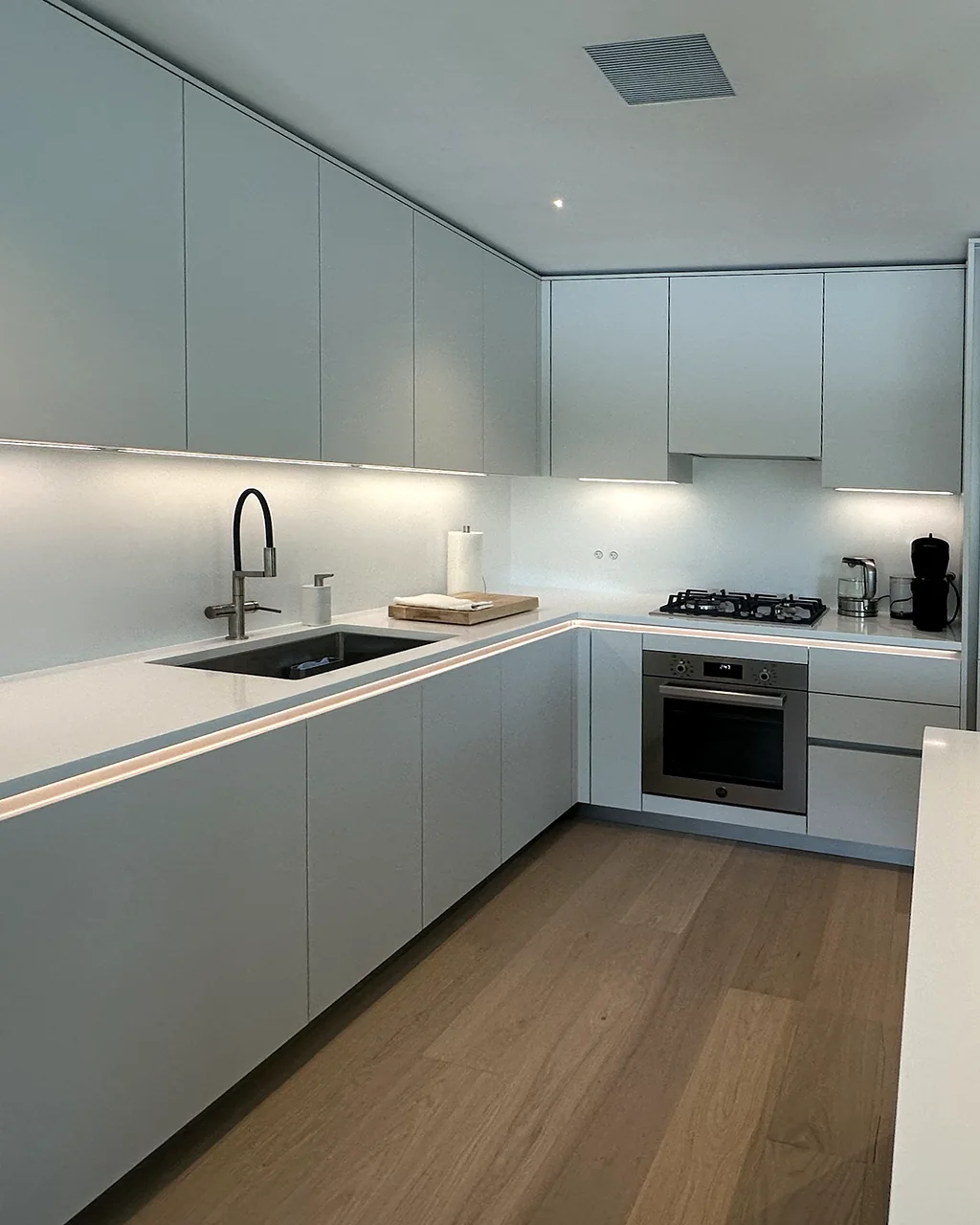 Modern white kitchen corner with a gooseneck faucet, gas cooktop, wall oven and under-cabinet lighting