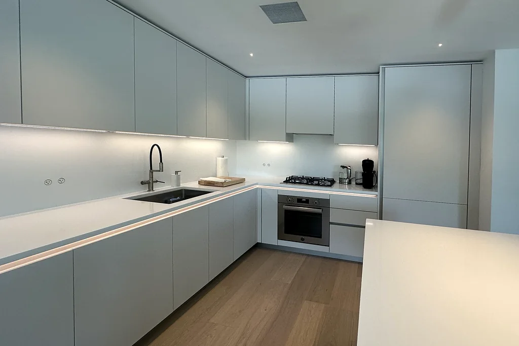 Modern L-shaped white kitchen with handle-free cabinets, under-cabinet lighting, a gas cooktop and a quartz island