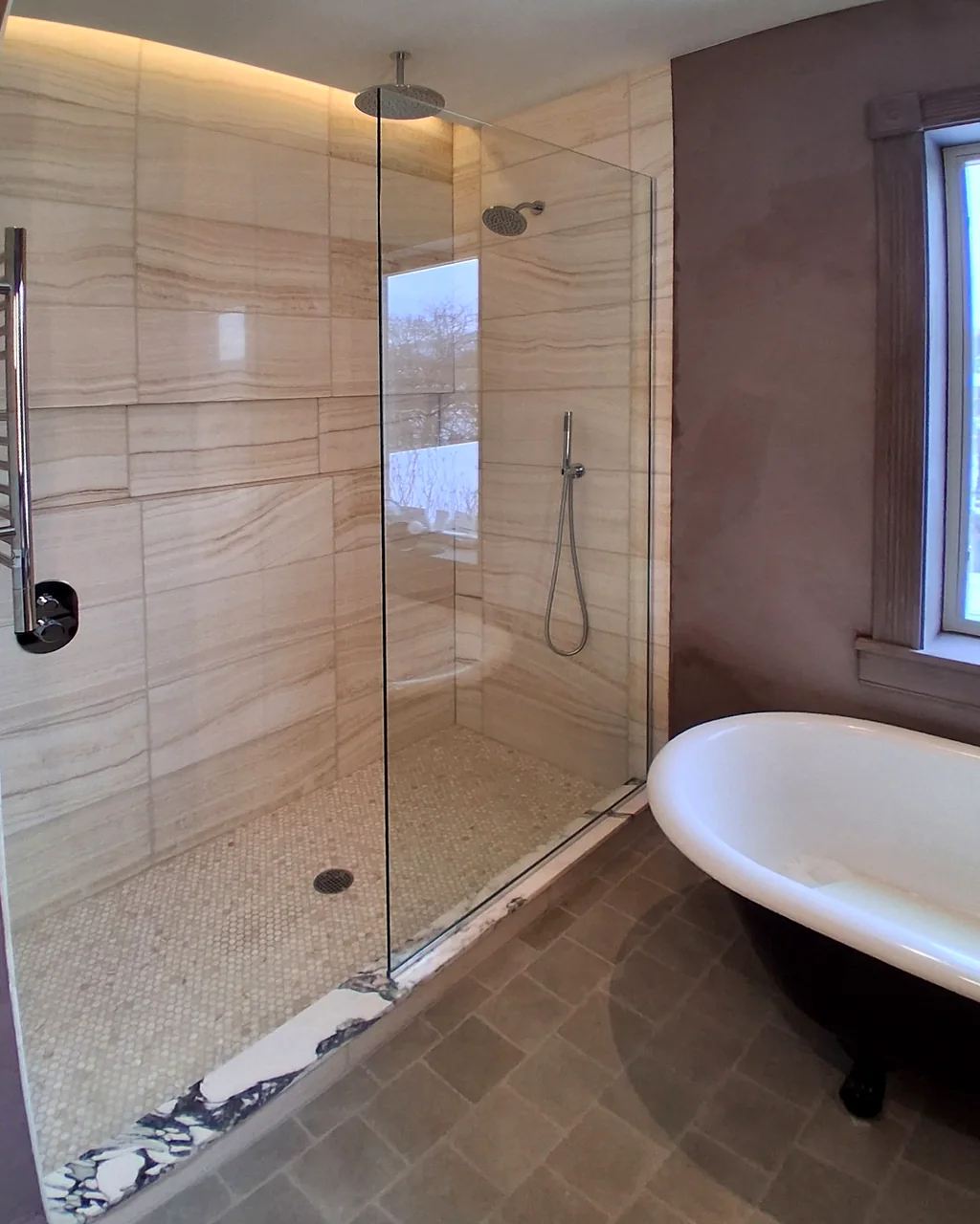 Walk-in shower with large stone-look wall tile, a frameless glass panel and a handheld shower, next to a clawfoot tub