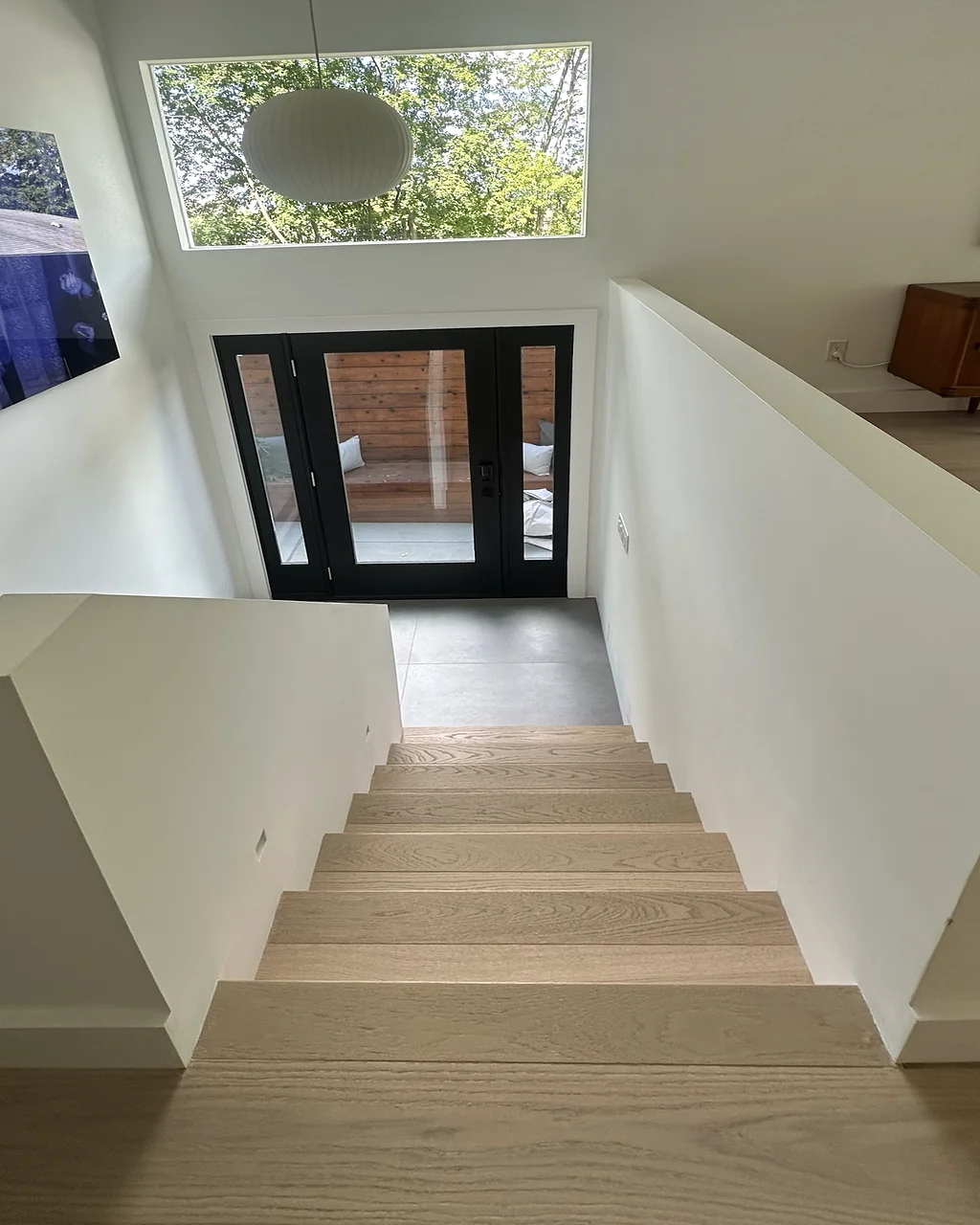 White oak stairs leading down to a black-framed glass entry door under a clerestory window