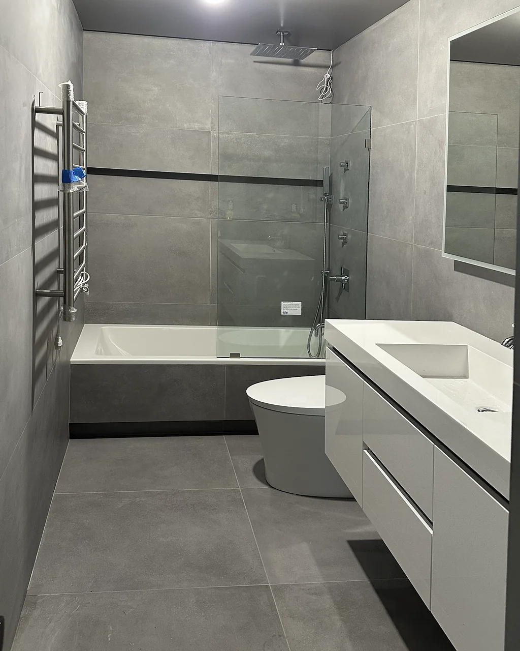 Floating white vanity, toilet and tub with a glass shower panel in a gray tile bathroom