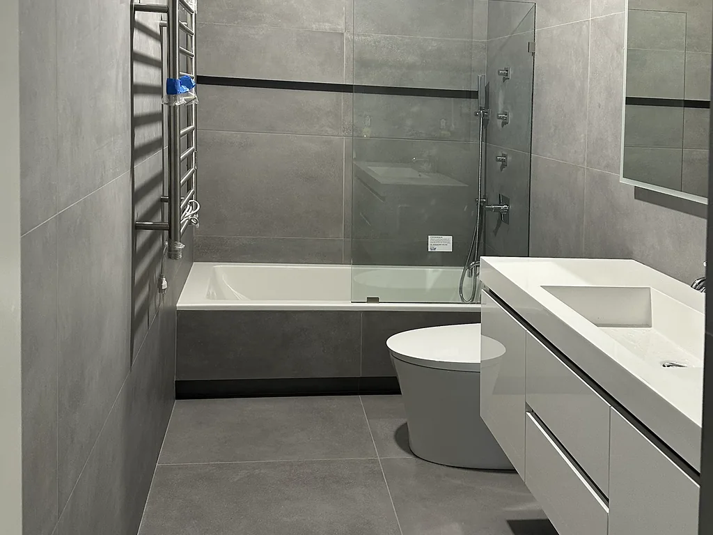 Floating white vanity, toilet and tub with a glass shower panel in a gray tile bathroom